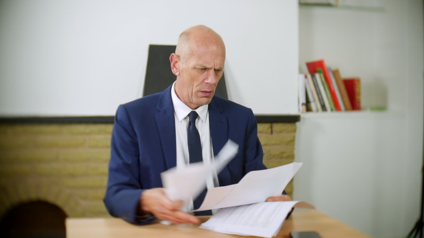 An angry  businessman sitting at a desk throws papers in the air. The man is in debt. The mature male shows his anger and desperation and holds his head in his hands. | Shutterstock HD Video #1097504067