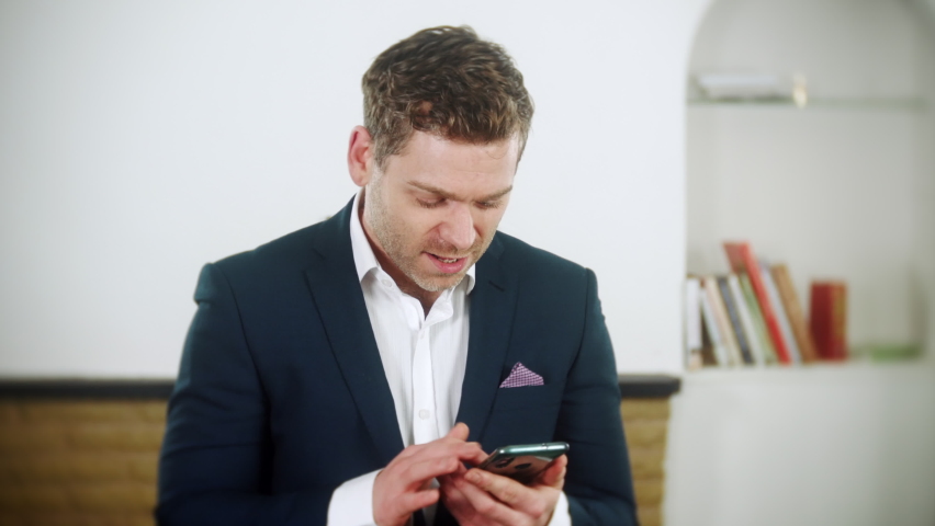 Young  businessman is using an app on his phone. He loses money and is upset. The man is wearing a blue suit and is at home in his living room. | Shutterstock HD Video #1097504071