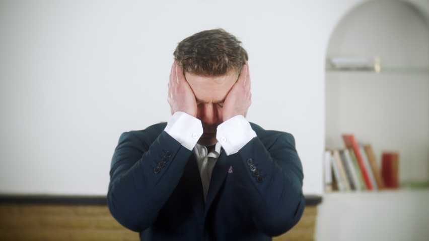 An entrepreneur businessman is distraught and depressed. He holds his head in his hands and looks lost and confused. The man is wearing a blue suit and is at home in his living room. | Shutterstock HD Video #1097504079