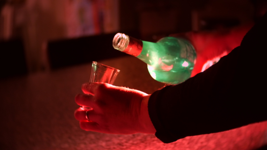 A shot of alcohol liquor is poured from a bottle into a glass. The drink is served at a bar in a nightclub with neon lights. | Shutterstock HD Video #1097504105