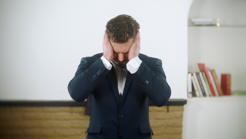 Young entrepreneur businessman is stressed and depressed. He holds his head in his hands. The man is wearing a blue suit and is at home in his living room. | Shutterstock HD Video #1097504109
