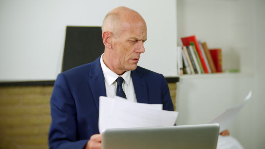 A mature businessman is confused and frustrated with working from home remotely. He is looking at paperwork while using a laptop computer. | Shutterstock HD Video #1097504111