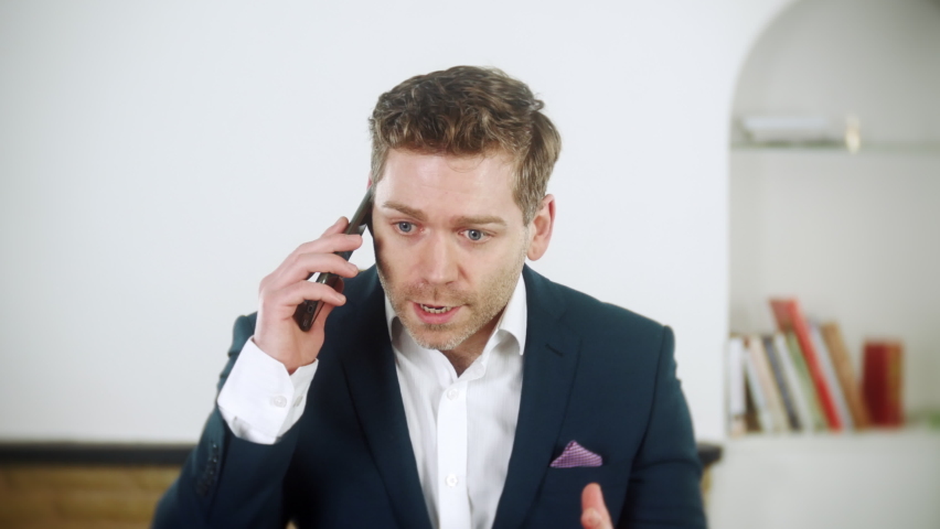Handsome young  businessman is shouting instructions on his phone. He is angry. The man is wearing a blue suit and is at home in his living room. | Shutterstock HD Video #1097504129