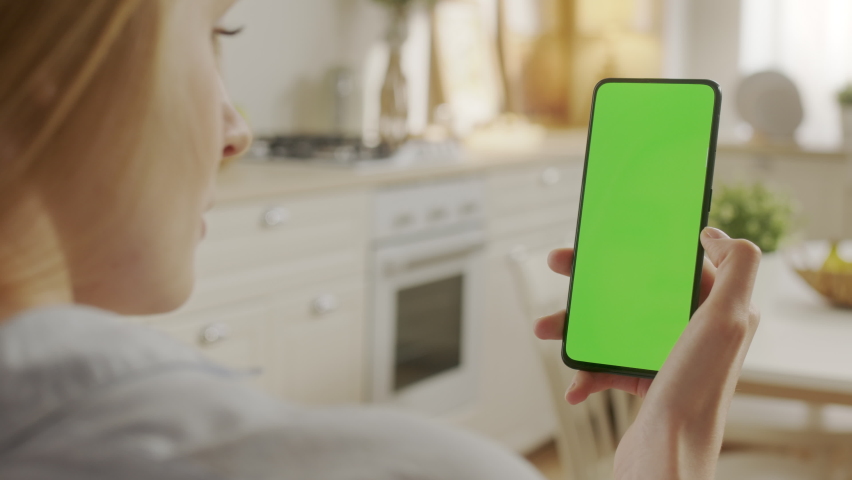 Handheld Camera: Back View of Woman at Kitchen Room Using Phone With Green Mock-up Screen Chroma Key Without Track Points Surfing Internet Watching Content Videos Blogs Delivery. Tapping on Center