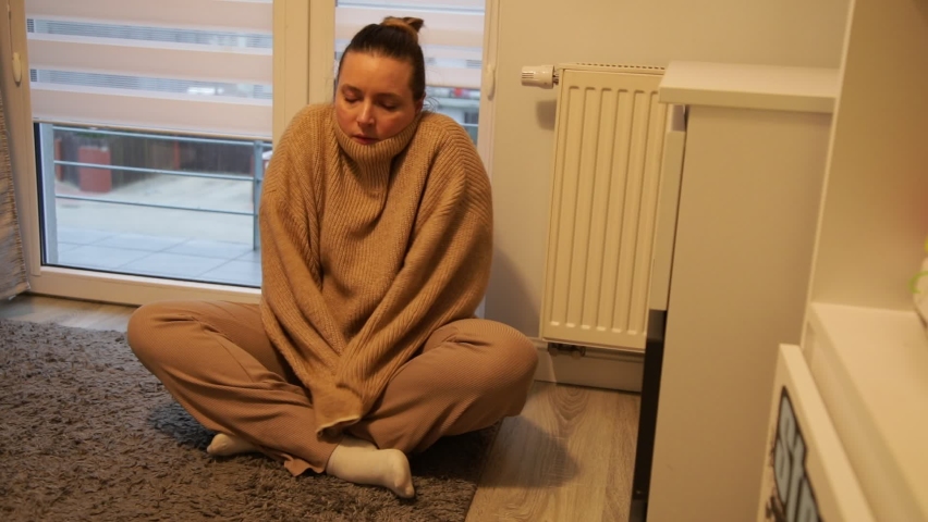 Frozen woman sit in cold room with dim light on floor. Wrap up body in warm white blanket and touch battery. Low temperature indoor. Winter, coldness, without heating, living conditions.