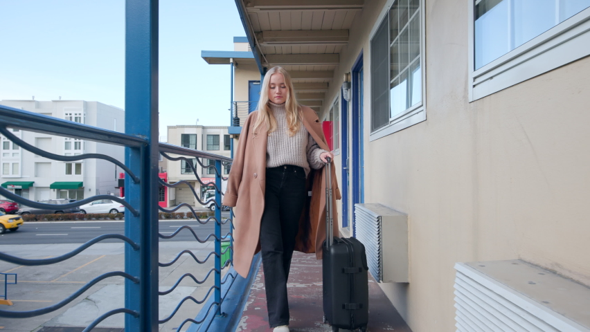 Caucasian, blonde girl strolling along motel corridor with a travel suitcase. Female traveler departing to the airport from a hotel complex. High quality 4k footage | Shutterstock HD Video #1097526217