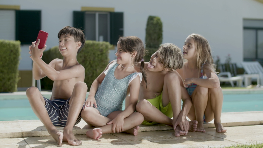 Happy children making selfie outside sitting on edge of pool. Boy holding mobile phone, taking photo. Children smiling and having fun. Handheld shot. Modern technologies, youth, friendship concept.