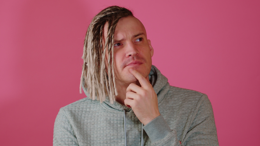 Young thoughtful handsome man with blonde dreadlocks on pink background. Pensive guy with doubt glance taps his chin with finger. Royalty-Free Stock Footage #1097529465