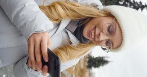 Vertical footage of a woman in eyeglasses typing by mobile phone outdoors. Closeup cheerful girl walking with smartphone in urban background. Smiling lady holding cellphone in hands outside. 4k video.