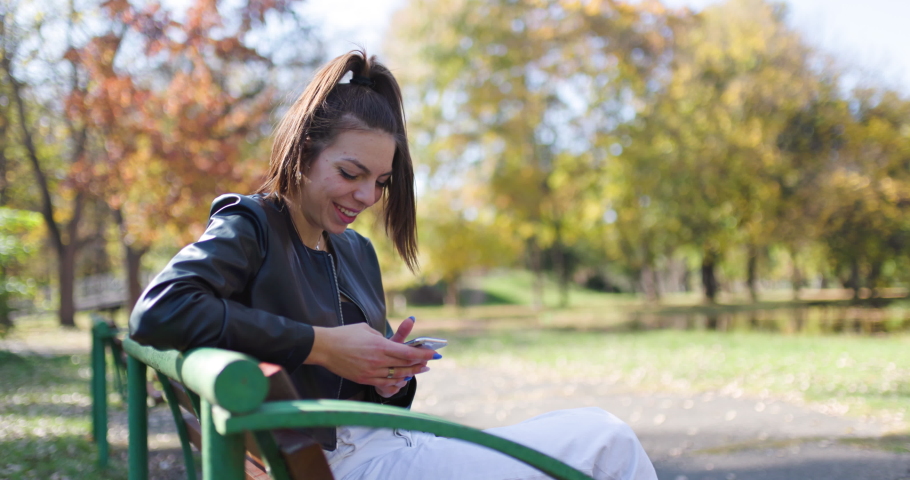 Beautiful girl sitting on a bench in the park. Fashionable young lady taking selfy photo with her phone.