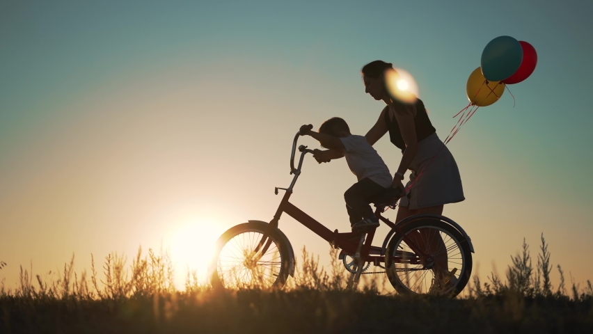 Happy family concept. KId learn to ride bike in park on green grass. Happy kid on his birthday. Mom teaches her son to ride bicycle at sunset in park. Mom helping hand to child. Boy on a bike in park Royalty-Free Stock Footage #1097536053