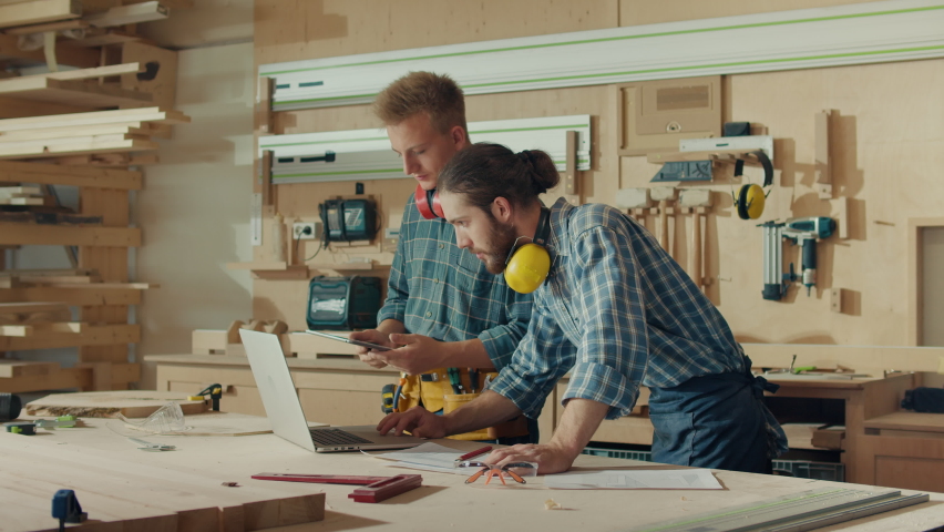 Small Business Owners of a Furniture Workshop Using Tablet, Notebook and Discussing the Design of a New Wooden Product. Handsome Carpenter and Young Male Apprentice Working in Studio. Royalty-Free Stock Footage #1097542659