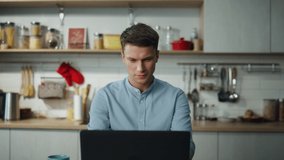 Portrait of attractive man browsing social networks at laptop sitting in home kitchen. Relaxed young guy messaging online with friends watching video searching internet. Freelancer working close up.