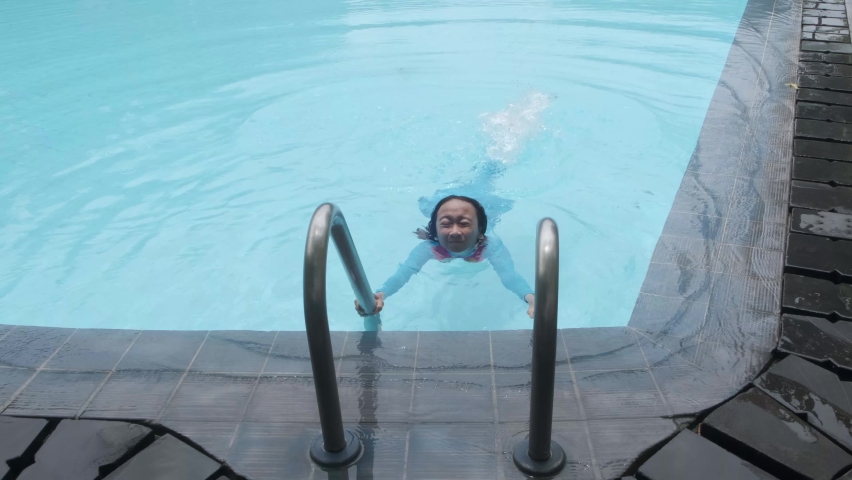 A girl learning to swim flapping her legs while holding on to a pool ladder