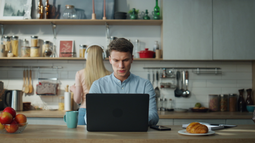 Serious busy manager have online zoom meeting with clients on laptop at kitchen while wife cooking breakfast. Young attractive businessman speaking on video conference at home. Remote work concept. | Shutterstock HD Video #1097553473