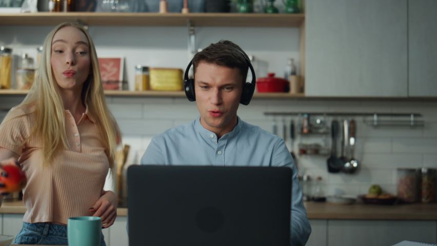Confident manager consulting online on web camera wearing wireless headphones sitting at kitchen while young blond wife dancing on backdrop. Attractive remote worker speaking on video conference. | Shutterstock HD Video #1097553475