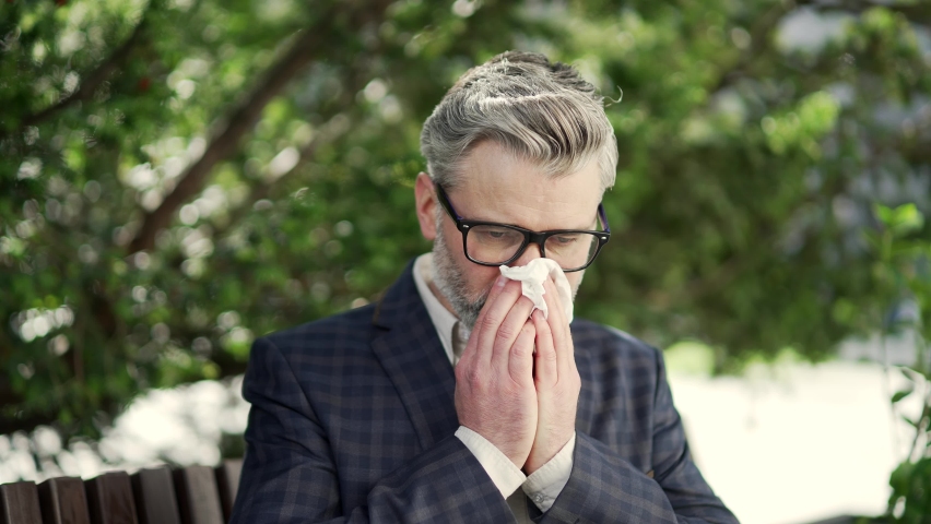 Close up of mature businessman suffers from allergies sneezes with a runny nose sitting on bench near business centre Unhealthy employee with glasses feeling unwell coughs a cold or flu outdoors alone | Shutterstock HD Video #1097557179