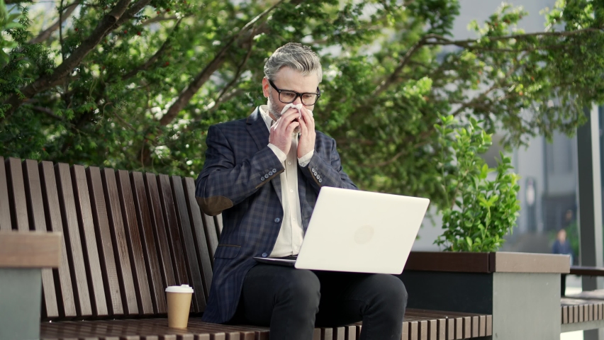 Handsome mature businessman suffers from allergies sneezes with a runny nose sitting on bench near business centre Unhealthy employee with laptop computer feeling unwell coughs a cold or flu outdoors | Shutterstock HD Video #1097557269