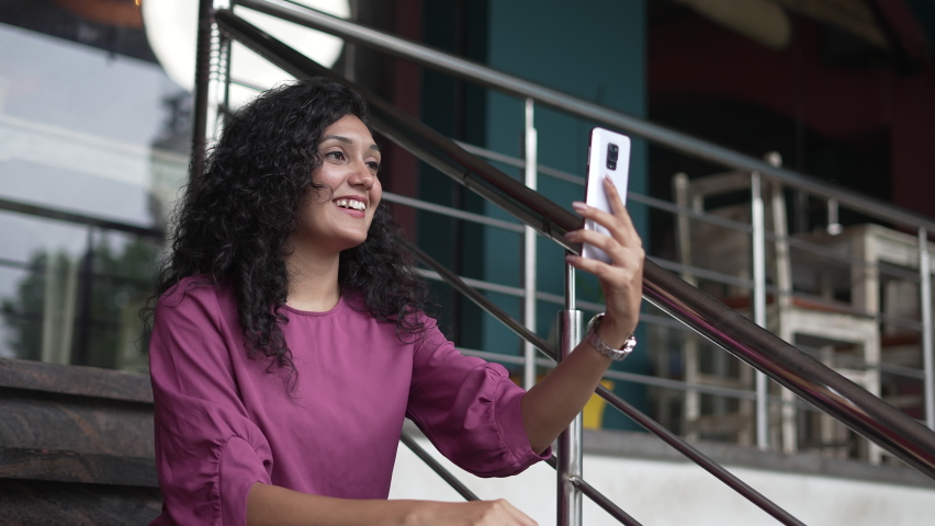 young Indian women holding smart phone doing videos call to close friend or social media content while siting in stairs.Smiling Asian female Conversation to friend and looking at mobile screen.