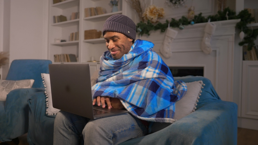 Black man in a blanket freezing indoors because of energy crisis, working indoors with a laptop in winter without central heating because of huge gas bills. High quality 4k footage | Shutterstock HD Video #1097564703