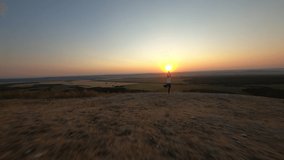 Cinematic FPV video of young woman doing yoga standing on one leg, arms outstretched on top mountain from which beautiful summer landscape, on background of bright sunlight in evening during sunset.
