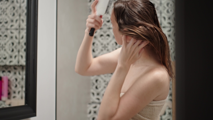 Wman wrapped in towel combs hair with hairbrush and dries it with hairdryer standing in bathroom. Women's morning hygiene and hair care Royalty-Free Stock Footage #1097565041