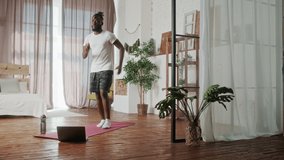Young Beginer African American handsome man doing cardio exercise in living room at home. Attractive active male doing workout follow instruction video from online trainer on laptop computer in house.