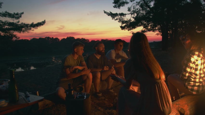 Friends sit on logs opposite each other on the banks of the river at sunset and laughingly communicate. Slow motion