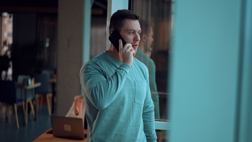 Fit mid-aged man in casual clothes stands at the window. Man looks into window and laughs speaking on the phone. Blurred backdrop. | Shutterstock HD Video #1097578165