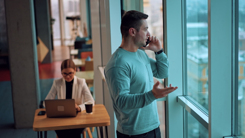 Dark-haired fit man stands at the window holding his phone at ear. Male listens to the speaker on the phone gesturing with his hand. Lady sits at desk at backdrop. | Shutterstock HD Video #1097578169