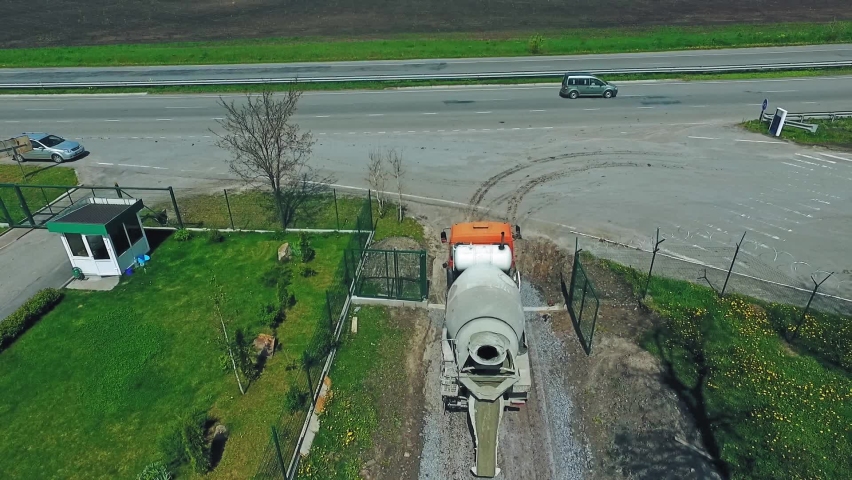 Concrete mixing machine moving by the road covered with gravel. Worker in vest and helmet walking next to the car. Highway and agricultural field at backdrop. | Shutterstock HD Video #1097578229