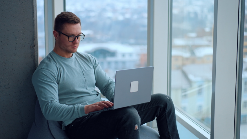Handsome fit man wearing glasses sits in a soft chair near the panoramic window. Man finishes his work, closes laptop and takes off his glasses leaning on the wall. | Shutterstock HD Video #1097578245