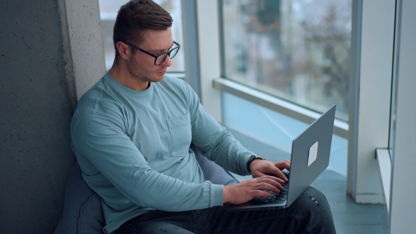 Thoughtful young mature man wearing glasses focused on the work at laptop. Man sits in bean bag chair leaned on the wall typing on the keyboard and glancing into window. Royalty-Free Stock Footage #1097578247