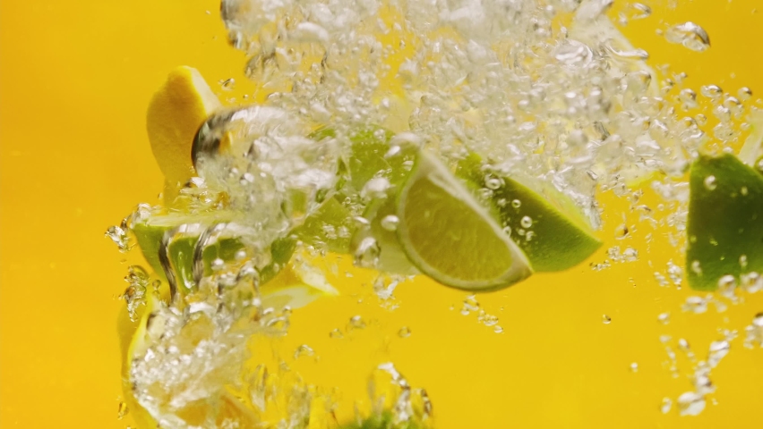 Close-up of falling sliced limes and lemons into the water on orange background, making a cocktail of citrus fruits, drinking cold lemonade, shooting of carbonated water with sliced fruits.  Royalty-Free Stock Footage #1097578329