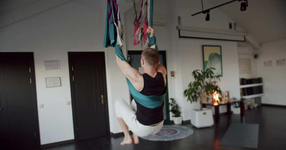 Man hanging upside down on aerial sinks. Pan around tilt down handheld shot of strong male hanging upside down on aerial hammock and bending body during yoga session in light studio Royalty-Free Stock Footage #1097580977