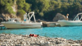 Red flip flop or sandal shoes on small pebbles beach, blurred calm turquoise sea with motor boats and trees behind. Afternoon sunny holiday resort scene - Powered by Shutterstock - Get 15% off with code: PIKWIZARD15