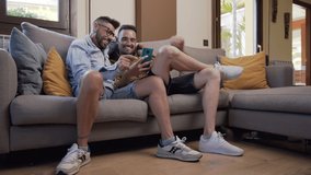 Happy young gay couple using mobile phone while sitting on a sofa in the living room. High quality 4k footage