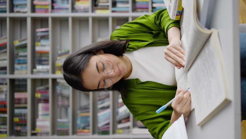 Young teacher in glasses writes lesson outline selecting exercises from various textbooks. Asian woman prepares for classes in library on blurred background Royalty-Free Stock Footage #1097589679
