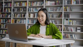 Smiling young teacher communicates with student by video call from library. Positive Asian woman shows study papers and explains material expressively
