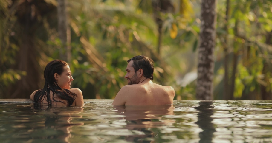 Young adult couple in love swimming together in open-air infinity pool enjoying tropical vacation on villa looking away at rainforest on sunset. Boyfriend and his girlfriend stand leaning on the pool | Shutterstock HD Video #1097593823