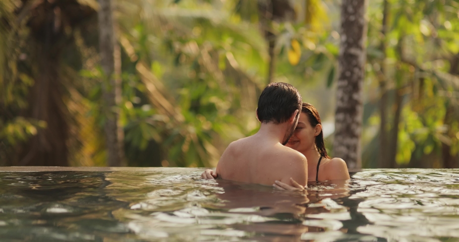 Young adult couple in love swimming together in open-air infinity pool enjoying tropical vacation on villa looking to each other against rainforest on sunset. High quality 4k footage Royalty-Free Stock Footage #1097593825