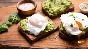 Avocado toast with poached egg. Cutting poached egg with a knife, tasty footage of runny egg yolk over mashed avocado on toasted rye bread, closeup view