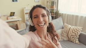 Handheld POV shot of cheerful young Caucasian woman in wireless headphones recording video blog or having video chat with friend during leisure time at home