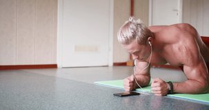 Young muscle shirtless man does plank exercises and listens to music through an online smartphone application. Sport and active lifestyle concept. 4k footage.