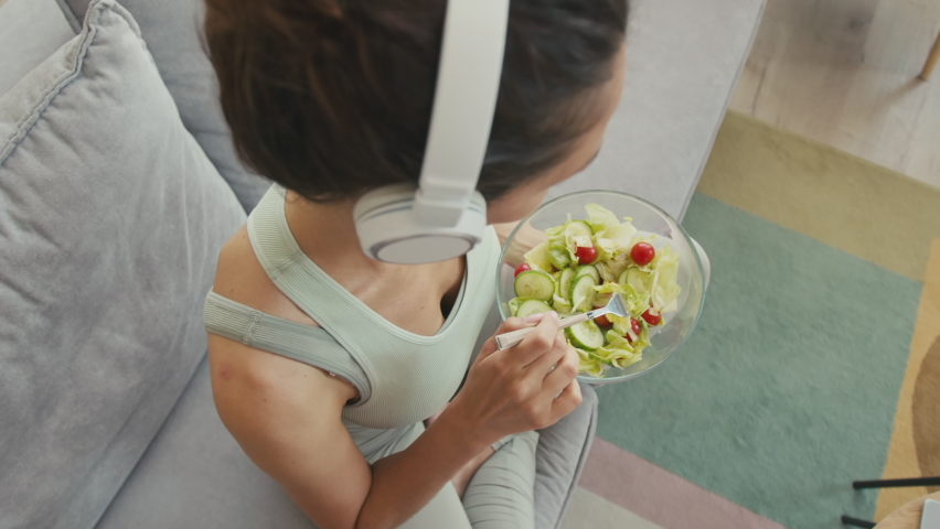 From above of young slim woman in wireless headphones and activewear eating healthy light green salad for lunch after sports training at home