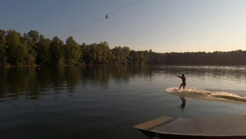 Aerial panning with drone over extreme female wakeboard surfer wearing helmet riding on lake water at cable wake park on background of bright sunlight at summer sunny day. Shooting in slow motion.