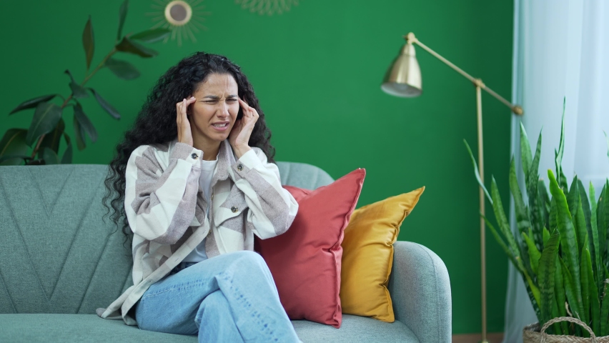Unhappy young female sitting on couch rubs temple with closing eyes suffering headache painful feelings chronic migraine high or low pressure Stressed upset dark haired woman feels hurt at home alone | Shutterstock HD Video #1097607003