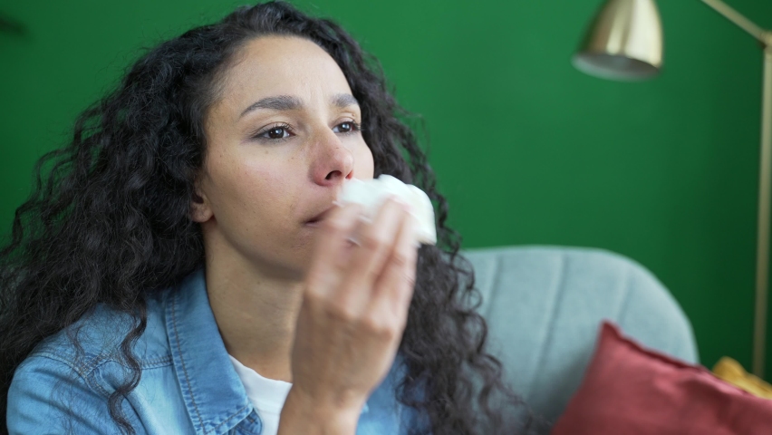 Close up portrait of young woman suffers from allergies sneezes with a runny nose sitting on sofa at home Unhealthy dark haired curly female feeling bad unwell coughs a cold or flu indoors alone  | Shutterstock HD Video #1097607061