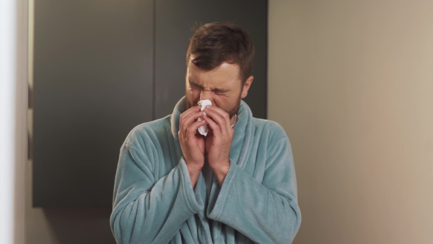 Portrait of a young man in a bathrobe sneezes and wipes his nose on a napkin | Shutterstock HD Video #1097611081