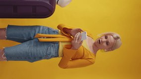 Vertical video. Hot tour. Excited woman. Vacation journey. Pretty surprised middle-aged lady expressing joy looking smartphone staying with suitcase on yellow background.